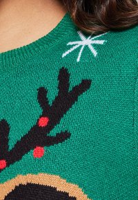 Grüner Strickpullover mit Rentiermotiv, schwarzen Geweihen, roten Akzenten und einem weißen Sterndetail am Ausschnitt.