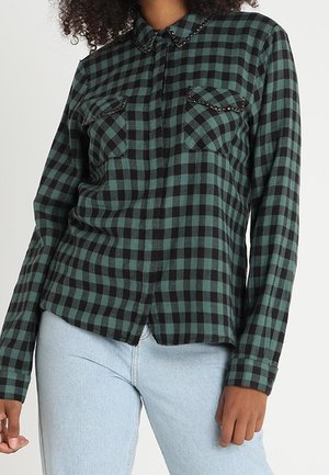 Femme portant une chemise à carreaux verte et noire à manches longues avec col clouté et poches, associée à un jean bleu clair.