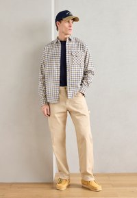 Baldai su patikrinimu beige ir mėlynos spalvos marškiniai virš tamsiai mėlynos spalvos marškinėlių, derinami su beige kelnėmis ir geltonais loafers, neutralios spalvos fone.