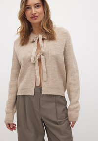 Bežna pletena cardigan z vezanjem spredaj in rebrasto teksturo. Ima dolge rokave s širokimi manšetami in skrajšano silhueto.