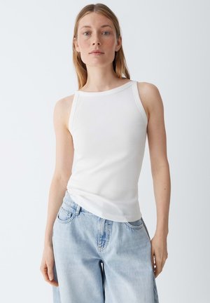 Vrouw met steil blond haar, gekleed in een mouwloos wit geribbeld topje en lichtblauwe high-waisted spijkerbroek, tegen een witte achtergrond.