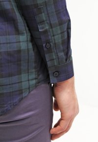 Mörkgrön och marinblå rutig skjorta med långa ärmar, knäppning vid ärmslut, mjukt tyg och avslappnad passform, visad på en persons arm.