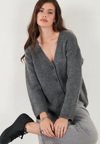 Cardigan gri tricotat cu decolteu în V, textură în relief, croială lejeră și patru nasturi pe față. Stilizat cu blugi gri și adidași negri.