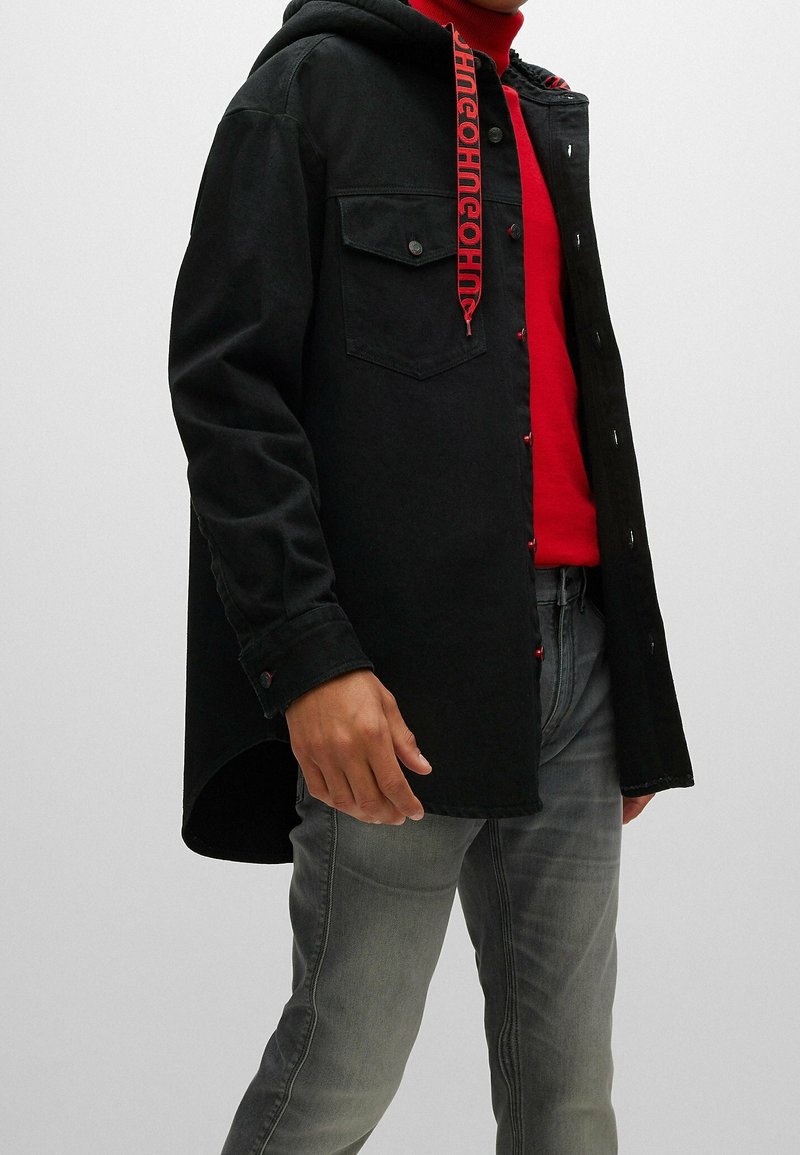 Personne portant une veste noire boutonnée avec des poches sur la poitrine, par-dessus un col roulé rouge et un jean gris délavé, debout devant un fond uni.