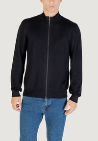 Hombre con una chaqueta negra de manga larga con cremallera y cuello alto, y jeans azules, de pie frente a un fondo claro y liso.