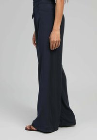 Pantalons large en velours marine, dotés d'une taille ajustée et de poches latérales, associés à des sandales ouvertes.