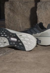 Ζευγάρι μαύρα και άσπρα αθλητικά παπούτσια Adidas, το ένα δείχνει την πατινάζ με σχέδιο, τοποθετημένα σε μια πέτρινη επιφάνεια με πέτρινους ογκόλιθους στο φόντο.