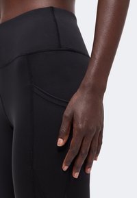 Gros plan d'une main à la peau foncée reposant à côté d'un legging de sport noir taille haute avec une petite poche latérale.