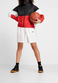 Röd, svart och vit färgblockerad jacka, vita shorts med svart Nike-logotyp, svarta träningssko, håller en basketboll.