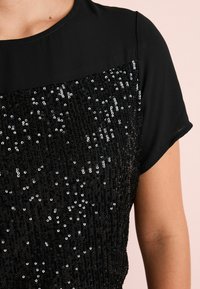 Top en sequins noirs avec un empiècement en tissu noir lisse, et un motif de sequins texturé et scintillant couvrant la partie inférieure. Manches courtes.