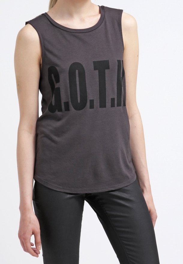 Haut sans manches gris foncé avec de grandes lettres noires "G.O.T.M." porté avec un pantalon en cuir noir par une personne debout.