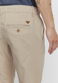 Beige Leinenhosen mit einer strukturierten Oberfläche, ausgestattet mit einem dreieckigen Lederetikett und einer geknöpften Gesäßtasche. Sichtbare Nähte.