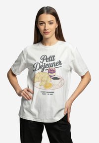 Balta kokvilnas T-kreklu ar brokastu grafiku, kurā attēlots kruasāns, kafija, ievārījums un apelsīna šķēle. Teksts ietver "Petit Déjeuner."