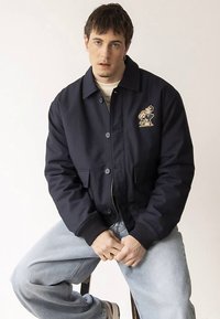 Tengerészkék bomberdzseki elülső zsebbel, két oldalzsebbel, gombos zárral és játékos hímzett mintával a bal mellrészen.