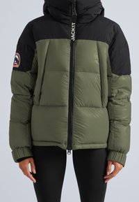 Person wearing a green and black padded jacket with a hood, front zipper, side pockets, and a logo patch on the left sleeve.