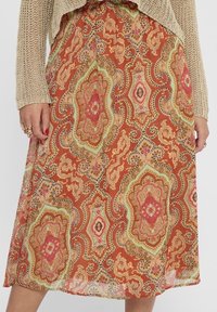 Une personne portant un pull en tricot beige tient une jupe midi colorée à motifs avec des designs élaborés dans des tons chauds.