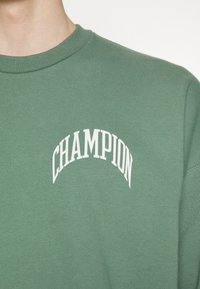 Zelená bavlněná mikina se žebrovanou strukturou a bílým logem "CHAMPION" ve tvaru oblouku na hrudi.