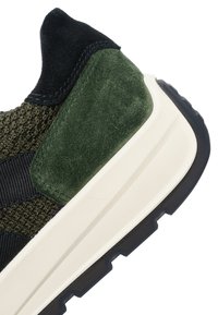 Primer plano del talón de una zapatilla con un panel de ante verde, parte superior de tela de punto negra, entresuela blanca y suela exterior de caucho negro con patrón de dibujo.