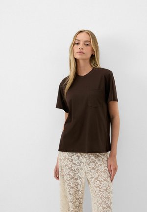 Młoda kobieta z długimi blond włosami nosi ciemnobrązowy T-shirt z krótkim rękawem oraz kremowe koronkowe spodnie, stoi na jednolitym białym tle.