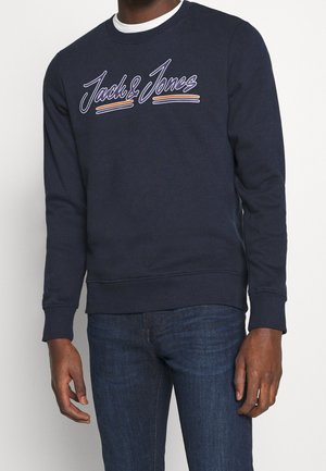Homme portant un sweat bleu marine avec le texte "Jack & Jones" et des rayures sur la poitrine, associé à un jean bleu foncé.