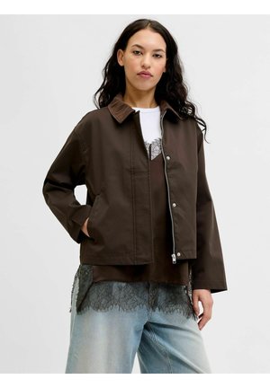 Jeune femme aux longs cheveux noirs porte une veste marron sur un top orné de dentelle et un jean bleu clair, debout devant un fond uni.