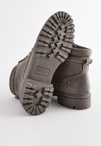 Botas de tobillo de ante marrón con una suela de goma resistente, que presenta surcos para tracción, etiquetadas como "NEXT" en la mediasuela y un collar doblado.
