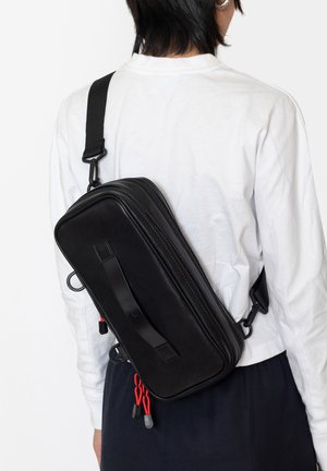 Sac bandoulière noir de forme rectangulaire, fabriqué en matériau lisse. Il dispose d'une sangle frontale, de fermetures éclair et de cordons d'accent rouge.