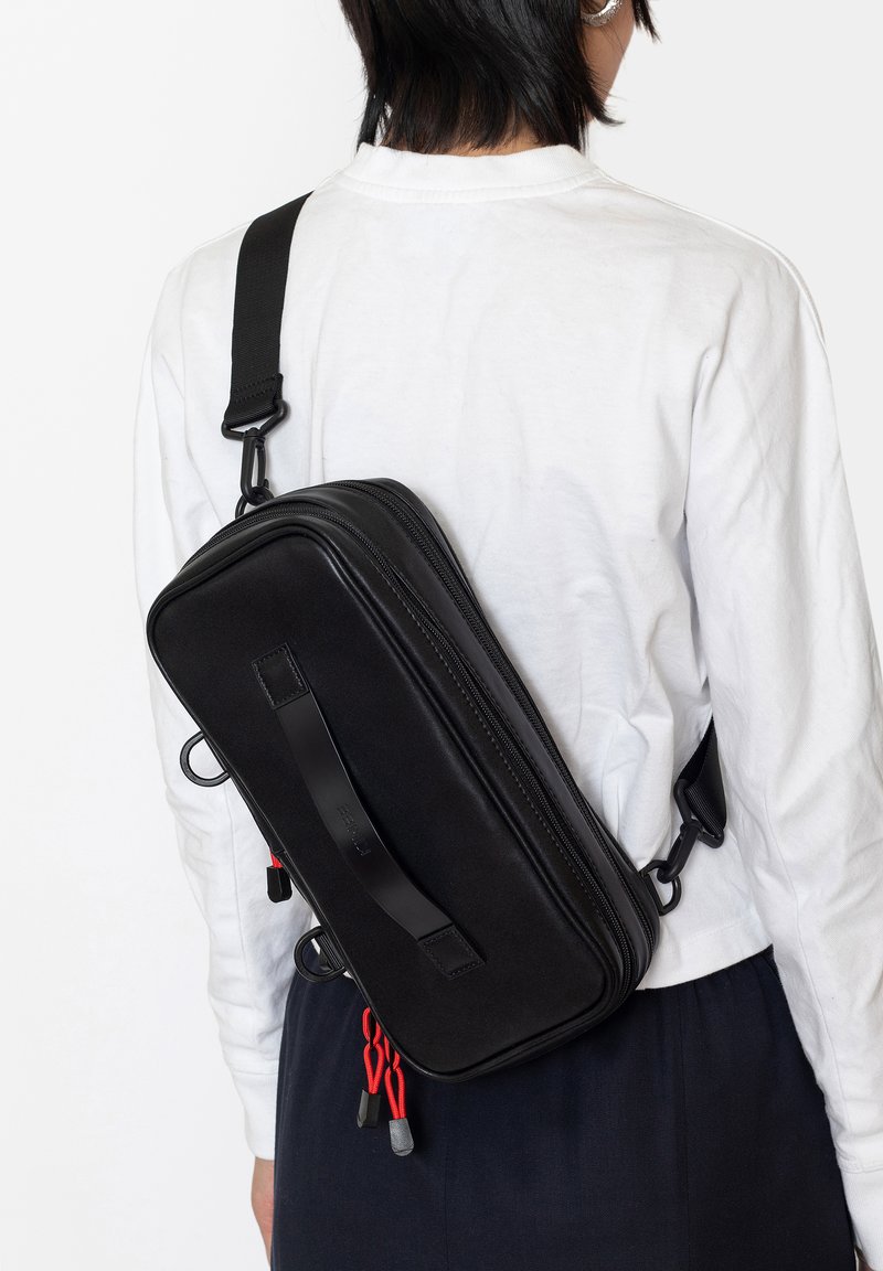 Borsa a tracolla nera di forma rettangolare, realizzata in materiale morbido. Presenta una tracolla frontale, cerniere e corde di accento rosse.
