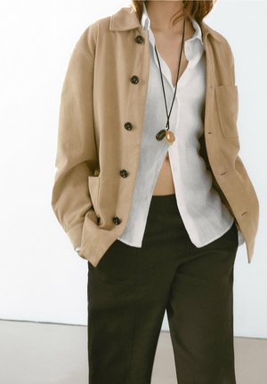 Personne portant une chemise blanche sous une veste beige boutonnée, un pantalon noir et un collier avec deux grands pendentifs ronds.