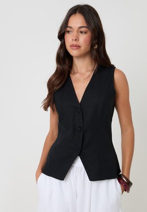 Kobieta o długich ciemnych włosach, ubrana w czarną kamizelkę na guziki bez rękawów oraz białe spodnie, z rękami w kieszeniach, na jednolitym tle.