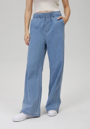 Femme portant un jean bleu clair à jambes larges avec taille élastique et baskets blanches, debout devant un fond uni gris clair.