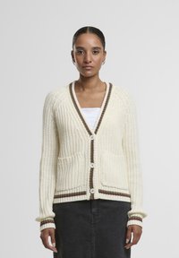 Off-white pleteni kardigan s V-izrezom, naglašen smešnim prugama, dva prednja džepa i kopčanje na dugmiće. Teksturalna tkanina i rebraste manžete.