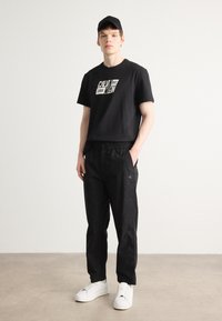 Fekete pamut póló fehér Calvin Klein logóval, fekete elasztikus derékú nadrággal és fehér sportcipőkkel párosítva, semleges háttér előtt.