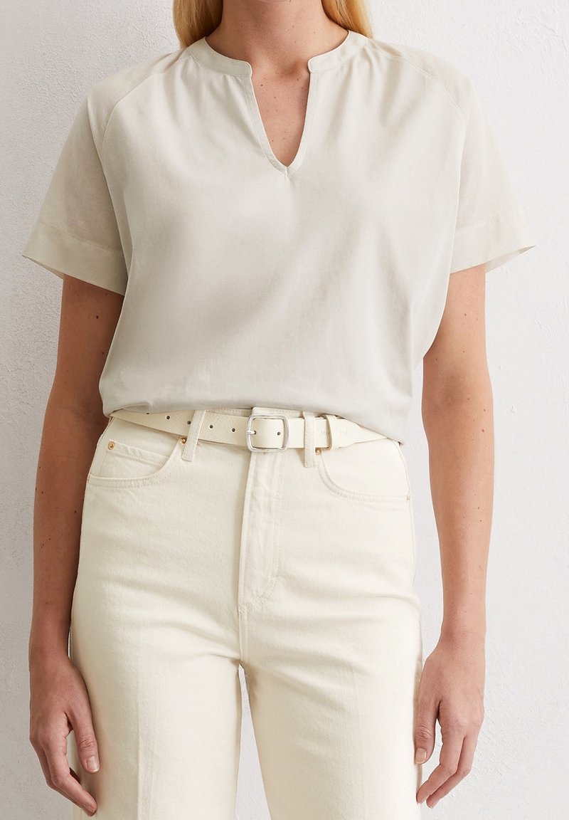 Femme portant un haut beige clair à manches courtes avec un col en V, rentré dans un jean crème taille haute avec une ceinture assortie.