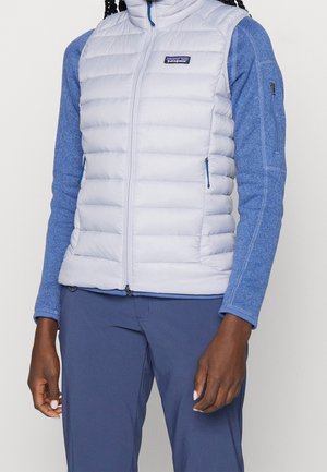 Person trägt eine hellgraue gesteppte Weste über einem blauem Fleece-Langarmshirt und dunkelblauer Hose, steht mit entspannten Armen an den Seiten.