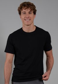 Black cotton t-shirt with short sleeves and a round neck, featuring a smooth texture and a fitted shape, worn by a smiling person.