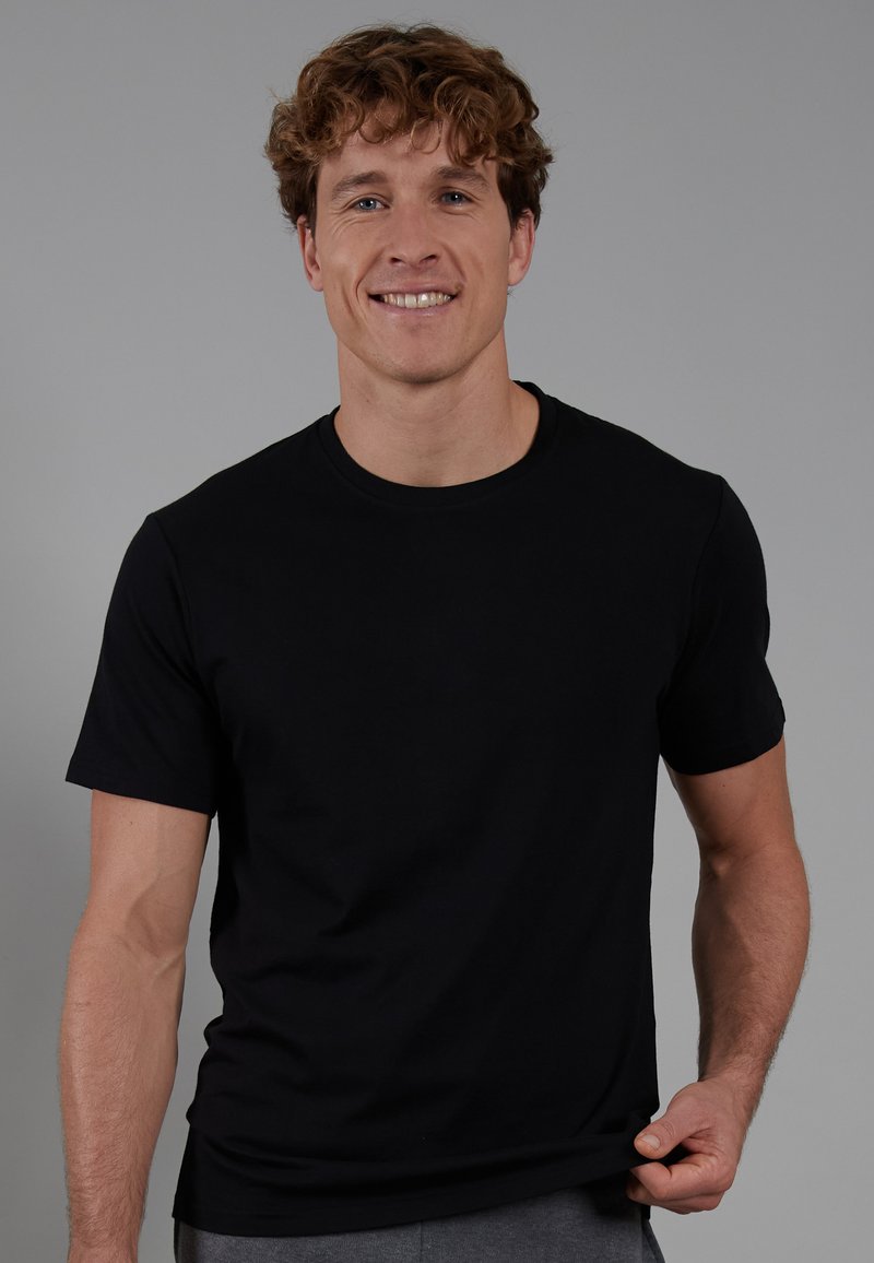 Black cotton t-shirt with short sleeves and a round neck, featuring a smooth texture and a fitted shape, worn by a smiling person.