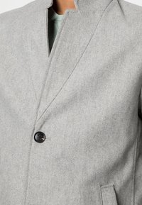 Cappotto a spina di pesce grigio chiaro chiuso con un bottone nero, indossato sopra una maglietta a girocollo verde chiaro.