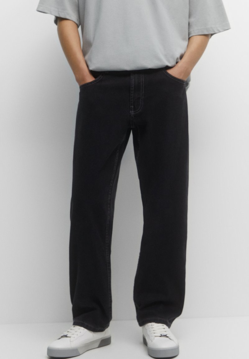 Personne portant un jean noir ample, une chemise à manches courtes gris clair, des baskets blanches, les mains dans les poches, debout devant un fond uni.