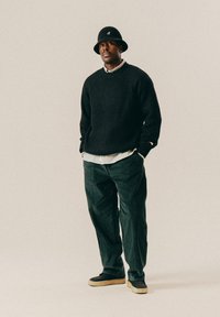 Mann mit schwarzem Bucket Hat, schwarzem Strickpullover, gestreiftem Hemd, dunkelgrüner Hose und schwarzen Schuhen mit beigen Sohlen, der mit den Händen in den Taschen steht.