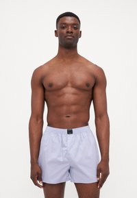 Pantaloni scurți de baie de culoare violet deschis, cu talie elastică și etichetă neagră. Textură fină, lungime până la genunchi, fără modele sau accente vizibile.