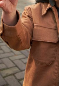 Bruine, textuurjacket met twee borstzakken, een ritssluiting en een col-nek. De stof lijkt duurzaam en stevig.