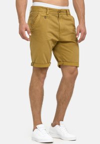 Shorts en toile jaune moutarde, avec des poignets roulés, une fermeture à bouton et des poches sur les côtés ; associés à des baskets blanches.
