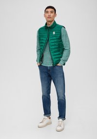 Grüne gepolsterte Weste mit vertikaler Steppstruktur über einem petrolfarbenen Pullover, kombiniert mit blauen Jeans und beigen Sneakers. Mit einem Logopatch.