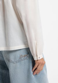Weiße, leichte Bluse mit langen Ärmeln und geknöpften Manschetten, kombiniert mit hellblauen Jeanshosen mit gestickten Details.
