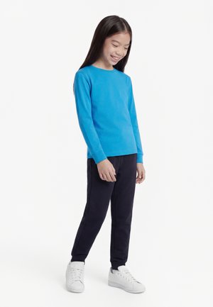 Ragazza sorridente con capelli lunghi e scuri, indossa una maglietta a maniche lunghe blu brillante, pantaloni neri e scarpe da ginnastica bianche, in piedi davanti a uno sfondo bianco.