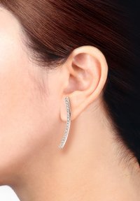 Silberne, geschwungene Ohrringe mit einer Reihe klarer, glitzernder Steine, an einem Ohr gegen einen neutralen Hintergrund präsentiert. Glatte Metalloberfläche.