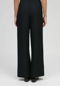 Personne portant un pantalon large à taille haute noir et des chaussures noires vernies, vue de dos sur un fond gris uni.