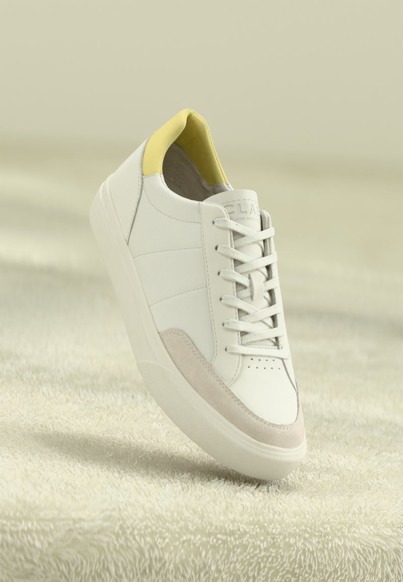 Weiße Leder Turnschuhe mit gelben Akzenten am Kragen, grauen Wildlederdetails an der Seite und einer strukturierten, weißen Gummisohle.
