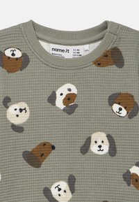 Camicia a maniche lunghe in tessuto oliva a nido d'ape con motivo faccia di cane in beige, marrone e nero. Collo rotondo a coste. Etichetta visibile.